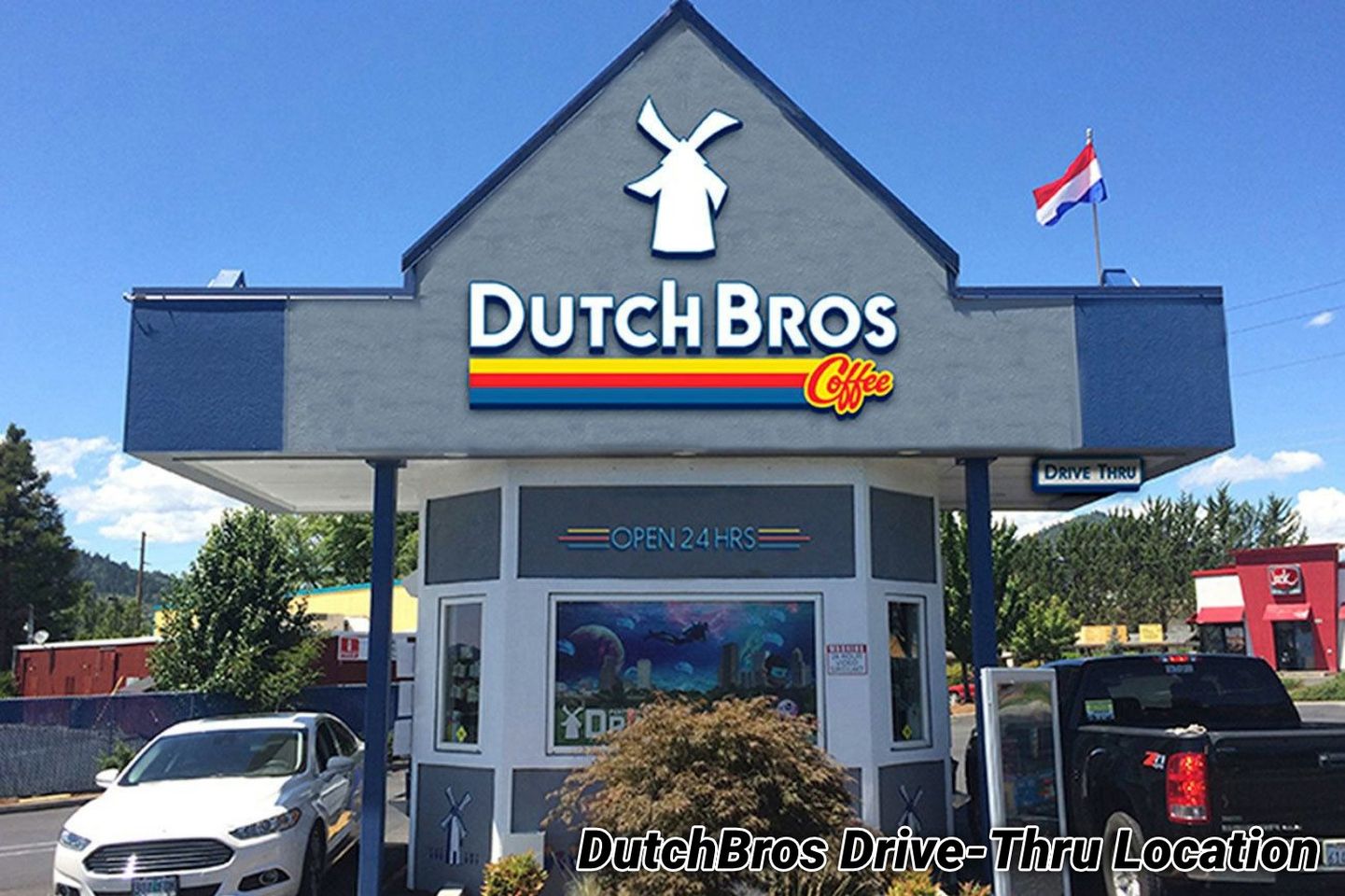 Exterior photo of an existing Dutch Bros Coffee drive-thru kiosk — a gray-and-blue stand with the windmill logo and an 'Open 24 Hrs' sign.